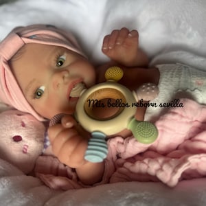 May include: A realistic baby doll with brown eyes, wearing a pink and white turban, is holding a wooden teether with a blue and green silicone design. The doll is lying on a white blanket with a pink blanket underneath.