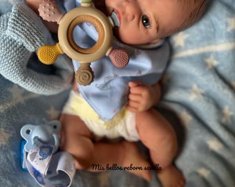 May include: A realistic reborn baby doll with pale skin, blue eyes, and fine brown hair. The doll wears a blue bib and a nappy. A multi-coloured teether is in its hand, and a dummy is in its mouth. The doll rests on a blue blanket.