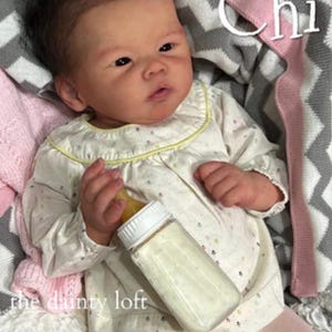 May include: A realistic baby doll with dark hair and a pink bow, wearing a cream-colored long-sleeve shirt with a small floral pattern and a yellow trim. The doll holds a clear plastic baby bottle with a white cap and a yellow nipple. The text Chi and the dainty loft are visible.