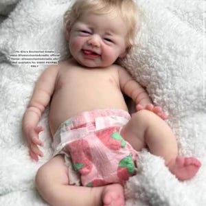 May include: A realistic reborn doll with blonde hair and a fair complexion, wearing a pink diaper featuring a strawberry pattern. The doll has a smiling expression with its tongue slightly visible. Text on the image reads: Fb: Ele's Enchanted Cradle, Insta: Eliesenchantedcradle official, Tiktok: eliesenchantedcradle, etc. Doll available for $0000 PAYPAL ONLY.