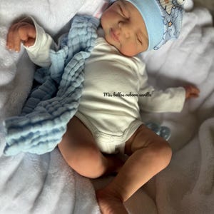 Puede incluir: Un bebé reborn durmiendo que lleva un body blanco, un gorro con estampado azul y blanco y una manta azul. El bebé tiene ojos marrones y una tez marrón claro. El bebé está acostado sobre una manta blanca y esponjosa. El texto "Mis bellos reborn sevilla" está impreso en el body.