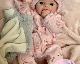 May include: A reborn baby doll wearing a pink onesie and matching hat with bunny ears. The outfit is decorated with floral and bunny prints. The doll is holding a gray stuffed animal and is lying on a pink blanket.
