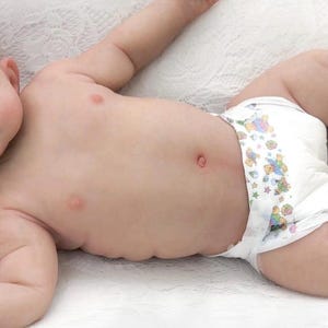 Może przedstawiać: Realistyczna lalka dla dziecka leży na białej powierzchni, ubrana w pieluchę z kolorowymi ilustracjami. Lalka ma niebieską opaskę na głowę z kwiatkiem i jest ułożona z wyciągniętymi ramionami. Oczy dziecka są zamknięte, co sugeruje sen.