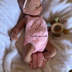 Puede incluir: Un conjunto de punto rosa con un dise&ntilde;o de ganchillo para una mu&ntilde;eca beb&eacute;. La mu&ntilde;eca lleva una diadema y tiene el pelo casta&ntilde;o. La mu&ntilde;eca est&aacute; tumbada sobre una manta blanca.