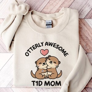 May include: Cream-colored sweatshirt with the text "Otterly Awesome T1D Mom" in black. The design features two cartoon otters hugging, with a pink heart above them. The sweatshirt is folded, with the sleeves visible.