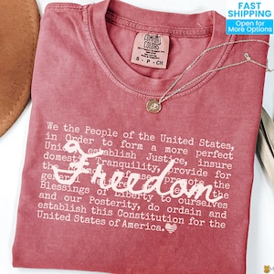 May include: A red Comfort Colors t-shirt with the text of the U.S. Constitution's preamble printed in white. A gold pendant necklace and a brown hat are also visible. The word "Freedom" is written in cursive.