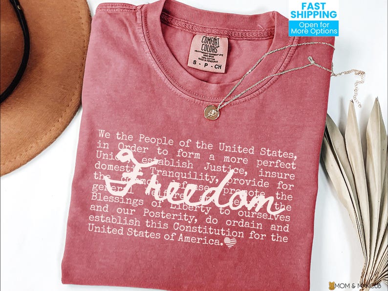 May include: A red Comfort Colors t-shirt with the text of the U.S. Constitution's preamble printed in white. A gold pendant necklace and a brown hat are also visible. The word "Freedom" is written in cursive.