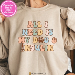 May include: A beige sweatshirt with the text "ALL I NEED IS MY DOG & INSULIN" in colorful letters. The text is in a retro font style. The sweatshirt has a crew neck and long sleeves.