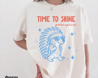 May include: An ivory t-shirt featuring a graphic of a porcupine wearing sunglasses and a backpack. The text above the porcupine reads "TIME TO SHINE" in orange and "porcupine" in cursive orange script. Blue star accents surround the porcupine.