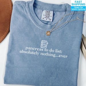 May include: A blue Comfort Colors t-shirt with the text "pancreas to do list: absolutely nothing...ever" in white. A gold necklace with a pendant is on the shirt. A brown hat is partially visible in the background.