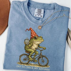 May include: A light blue Comfort Colors t-shirt featuring a whimsical illustration of a frog riding a bicycle and wearing a wizard hat. The frog holds a blood glucose meter displaying "300". The text "OFF TO CHECK MY KETONES" is printed below the frog.