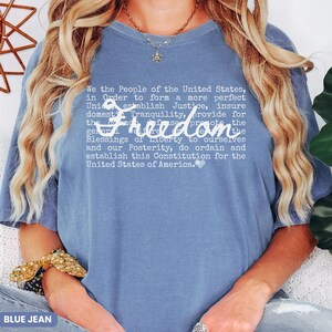 May include: A blue jean colored t-shirt with the word "Freedom" in a cursive font. The shirt features the text of the preamble to the United States Constitution in a white font.