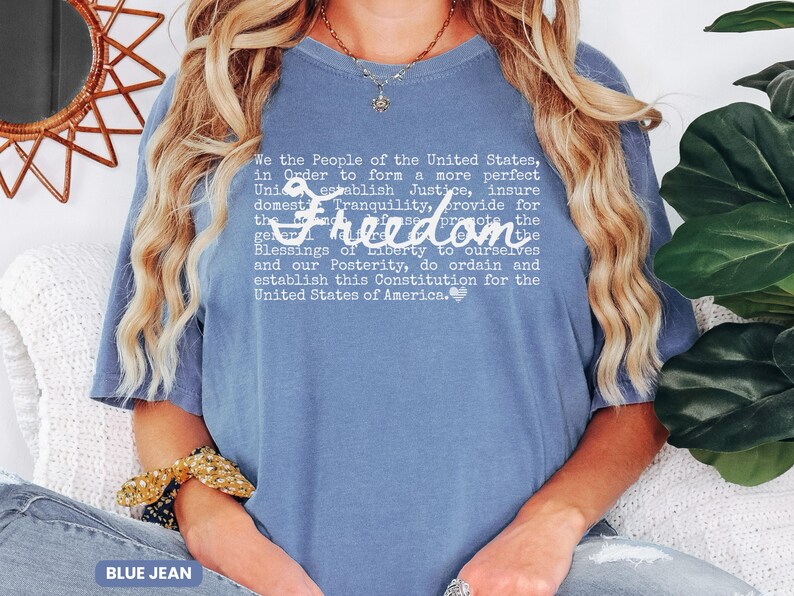 May include: A blue jean colored t-shirt with the word "Freedom" in a cursive font. The shirt features the text of the preamble to the United States Constitution in a white font.