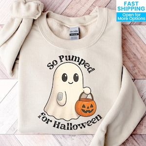 May include: A cream-colored crewneck sweatshirt featuring a graphic of a white ghost holding an orange jack-o'-lantern bucket. The text above and below the ghost reads "So Pumped for Halloween" in a dark, distressed font. The garment has a Gildan label.
