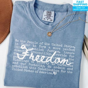 May include: A pale blue Comfort Colors t-shirt featuring the text "We the People...Freedom...United States of America" in white. A gold necklace with a pendant and a brown hat are also visible.