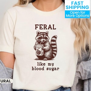 May include: White t-shirt featuring a brown raccoon illustration holding a juice box. The text "Feral like my blood sugar" is printed below the raccoon.