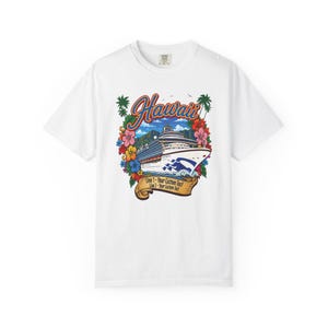 Puede incluir: Una camiseta blanca con un gráfico de un crucero navegando cerca de montañas, rodeado de coloridas flores de hibisco y palmeras. La palabra Hawaii está impresa en azul y naranja sobre el barco, con una cinta debajo para texto.