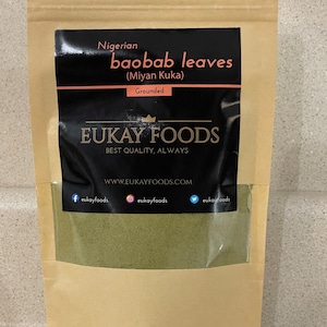 Peut inclure: Un sac en papier brun avec une fenêtre transparente montrant des feuilles de baobab vertes. L'étiquette noire indique "baobab leaves nigériens (Miyan Kuka) Grounded" et présente la marque "EUKAY FOODS". Le sac comprend également des réseaux sociaux.