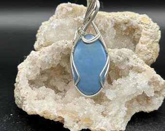 May include: A silver wire wrapped pendant featuring a light blue oval gemstone. The pendant is displayed on a white rock with a crystal-like texture.