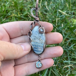 May include: A copper wire wrapped pendant featuring a blue and gray stone with a smaller blue stone dangling below.
