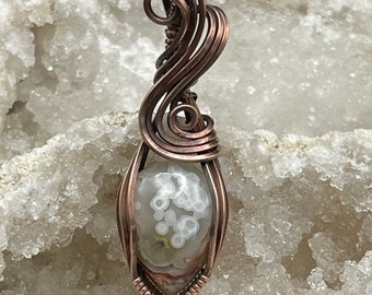 May include: A white and brown agate gemstone pendant wrapped in copper wire. The wire is twisted and curled into a decorative design.