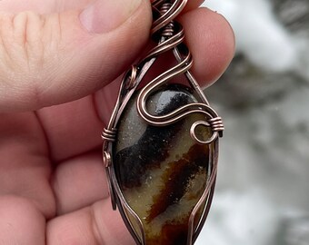 May include: A copper wire wrapped pendant featuring a brown and white agate stone. The pendant is shaped like a teardrop and has a delicate wire design.