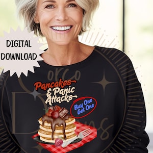 Può includere: Una felpa nera con una grafica di una pila di pancake con salsa al cioccolato e fragole. Il testo sulla grafica dice "Pancakes & Panic Attacks" e "Buy One Get One".