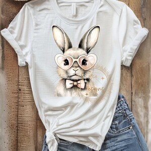 May include: White t-shirt featuring a watercolor illustration of a bunny wearing heart-shaped glasses and a bow tie. The bunny is surrounded by a gold design with the text "Arco Designs". The shirt is paired with blue denim jeans.