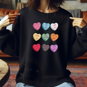 May include: A black sweatshirt with colorful heart-shaped candy conversation hearts printed on it. The hearts have various messages, including "Not Today, Cupid", "You Suck", "I Want Tacos", "Go On, Git", "Nah I'm Good", "Don't Be Mine", "It's Not Me It's You", "Where's The Tequila", and "Cuter Than U".