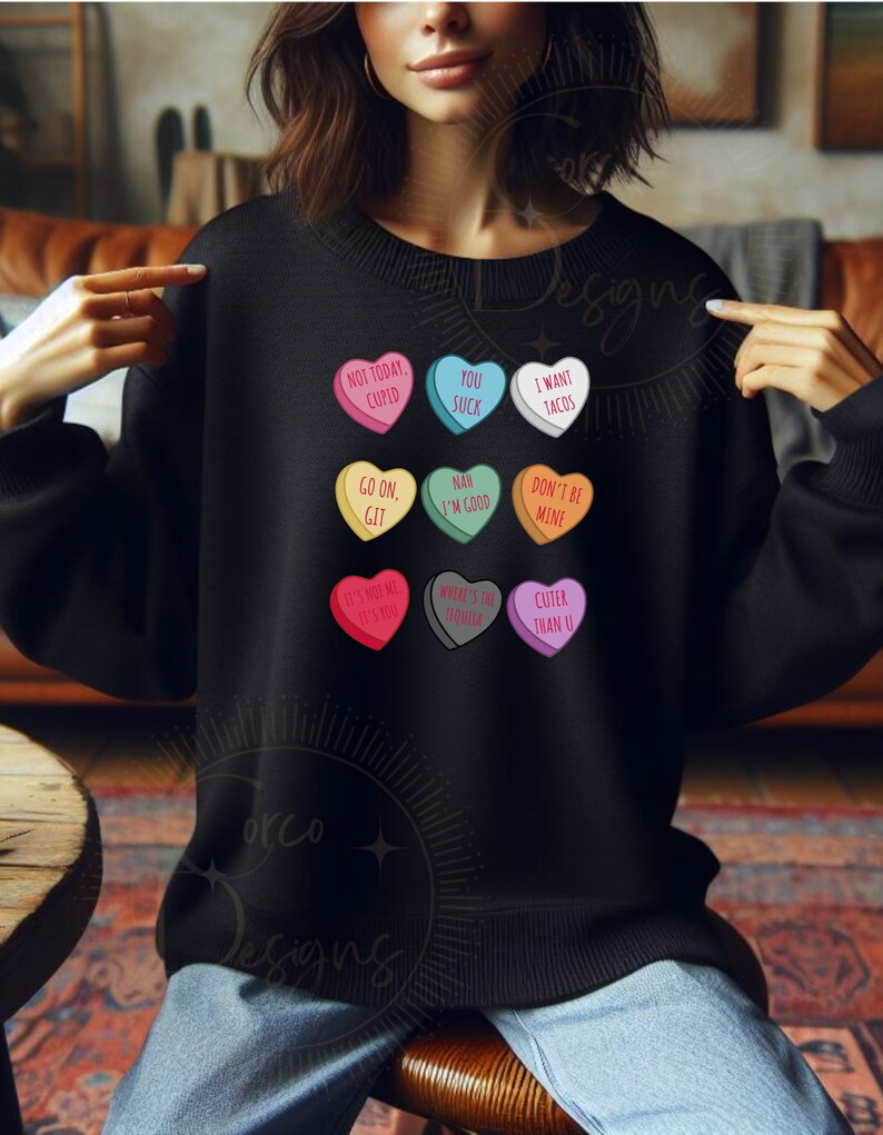 May include: A black sweatshirt with colorful heart-shaped candy conversation hearts printed on it. The hearts have various messages, including "Not Today, Cupid", "You Suck", "I Want Tacos", "Go On, Git", "Nah I'm Good", "Don't Be Mine", "It's Not Me It's You", "Where's The Tequila", and "Cuter Than U".