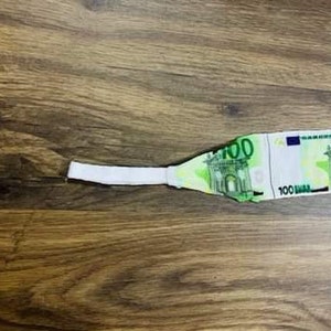 May include: A white fabric headband with a 100 Euro bill printed on it. The headband is lying on a wooden surface.