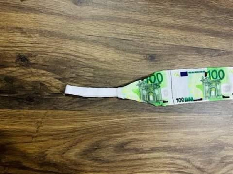 May include: A white fabric headband with a 100 Euro bill printed on it. The headband is lying on a wooden surface.