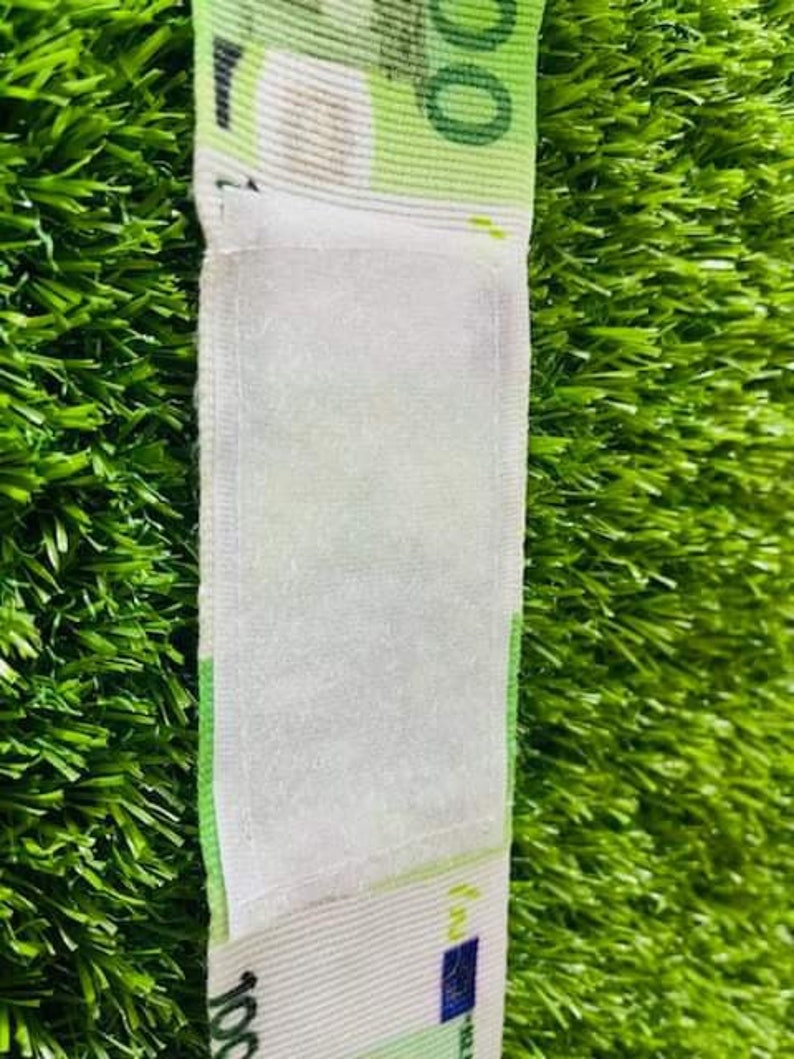 May include: A white fabric wristband with a euro bill design. The wristband has a white velcro closure.