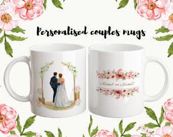 Op de afbeelding: Twee witte keramische mokken met gepersonaliseerde ontwerpen. Een mok heeft een aquarel-illustratie van een stel dat onder een bloemenboog staat. De andere mok heeft een bloemenontwerp met de tekst "Name1 en Name2".