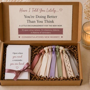 Puede incluir: Una caja de regalo de cartón marrón que contiene un juego de tarjetas blancas con el texto Open when you need a reminder you are loved, atadas con una cinta malva, y diez cintas de hitos en varios colores pastel, con un mensaje alentador.