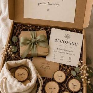 May include: A cardboard gift box containing a stack of envelopes tied with a sage green ribbon, a card titled BECOMING with 15 Open-When Letters, a small drawstring pouch, and several wooden discs engraved with words like breathe, move, nourish, begin, and grace.