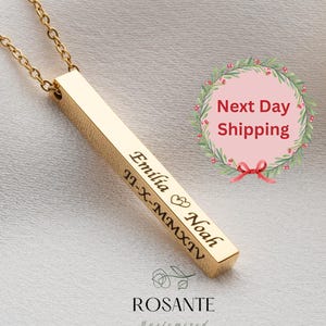 May include: A gold-colored bar pendant necklace with a delicate chain. The pendant is engraved with the names "Emilia" and "Noah", a heart symbol, and the date "11-X-MMXIV". The necklace is displayed on a white, textured surface.