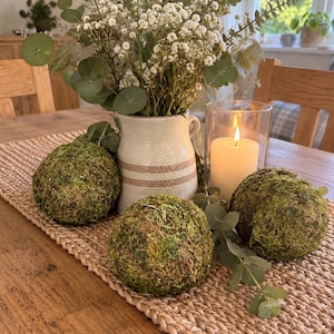 Pode incluir: Um centro de mesa composto por três bolas de musgo esféricas, um jarro de cerâmica com flores brancas de gipsofila e ramos de eucalipto, e uma vela de pilar acesa num suporte de vidro, sobre um caminho de mesa retangular entrançado.