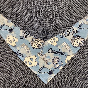 May include: Light blue pet bandana with a repeating pattern of UNC logos, Tar Heel mascots, and the words "North Carolina" and "Carolina". The bandana has white snap closures at the ends.