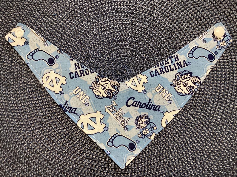 May include: Light blue pet bandana with a repeating pattern of UNC logos, Tar Heel mascots, and the words "North Carolina" and "Carolina". The bandana has white snap closures at the ends.