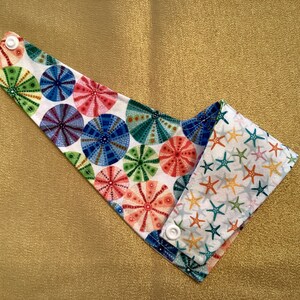 May include: A white fabric bib with a reversible design. One side features a colorful pattern of sea urchins, the other side features a white background with a pattern of colorful starfish.