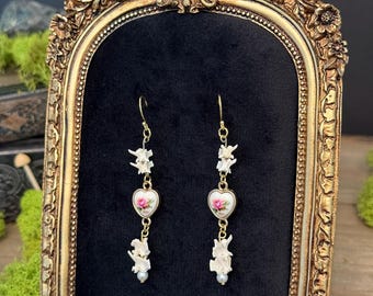 May include: A pair of gold-tone drop earrings featuring white bone-like segments, a central heart-shaped charm with a pink rose floral pattern, and a small white bead at the bottom, suspended from gold-tone ear wires.