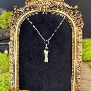 May include: A silver-toned chain necklace featuring a pendant with a small, off-white bone-shaped charm wrapped in thin silver wire, positioned below a single dark, round bead. The necklace is displayed against a black velvet background.