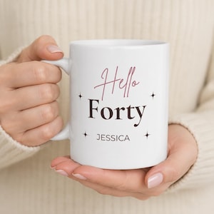 Könnte beinhalten: Ein weißer Keramikbecher mit dem Text Hello Forty in einer Kombination aus rosa Schreibschrift und dunkelbrauner Serifenschrift, darunter der Name JESSICA in einfacher serifenloser Schrift, verziert mit vier kleinen schwarzen Sternen.