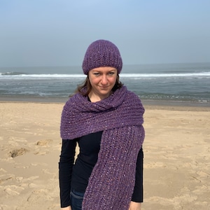 May include: A woman wearing a purple knitted hat and scarf on a sandy beach.