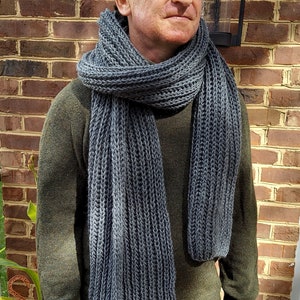 May include: A man wearing a dark gray knitted scarf. The scarf is long and has a ribbed texture. The man is wearing a green sweater and green pants.
