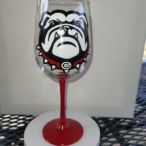 May include: A red wine glass with a hand-painted design of the University of Georgia bulldog mascot. The bulldog is white with black outlines and red eyes. The glass has a red stem and base.