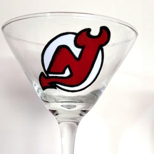 May include: A clear martini glass with a red and white logo of the New Jersey Devils hockey team. The logo is a stylized "N" and "J" inside a white circle, outlined in black.