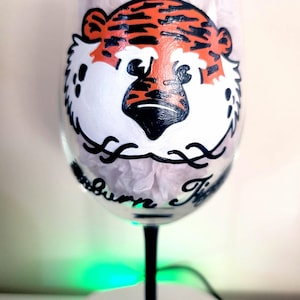 May include: A clear wine glass with a black stem and base. The glass is decorated with a hand-painted orange and black tiger face. The text "Auburn Tigers" is written in black cursive below the tiger.