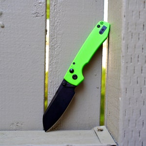 May include: A green and black pocket knife with a black blade and a green handle. The knife is partially inserted between two wooden slats.